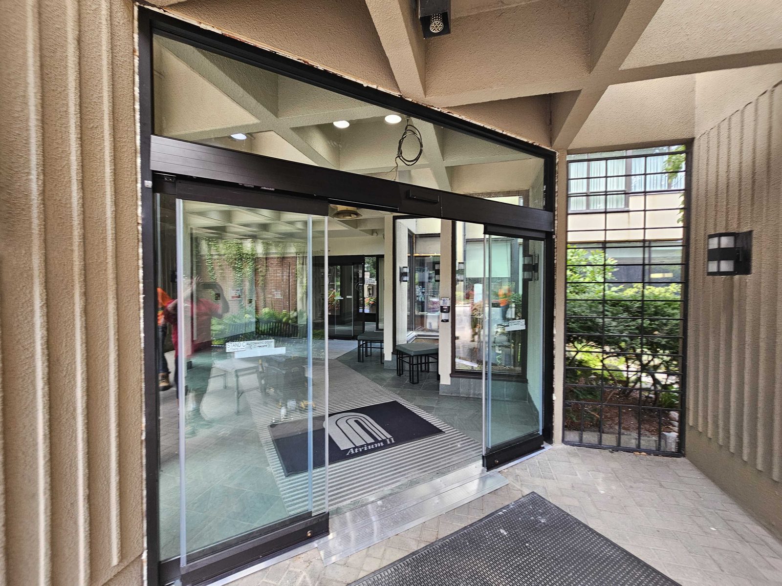 A bi-parting automatic sliding entrance with black aluminum framing and glass leaves standing open at a condominium lobby, a low threshold at floor level, an "Atrium II" entrance mat inside and a floor mat outside, under a concrete canopy