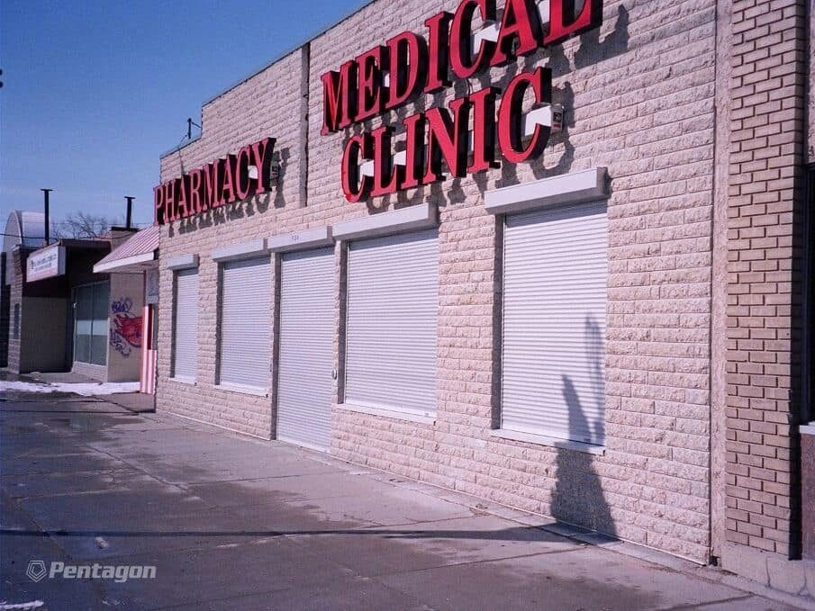 White aluminum rolling shutters closed over the window bays of a pharmacy and medical-clinic storefront — a Pentagon product photograph of the system we supply